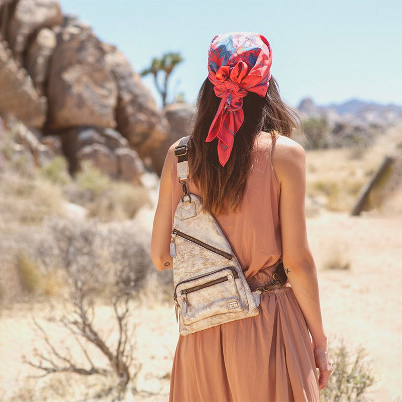 Woman with brown hair wearing a mauve dress, a floral bandana on her head and a cross body back pack in a dessert rock scene. 