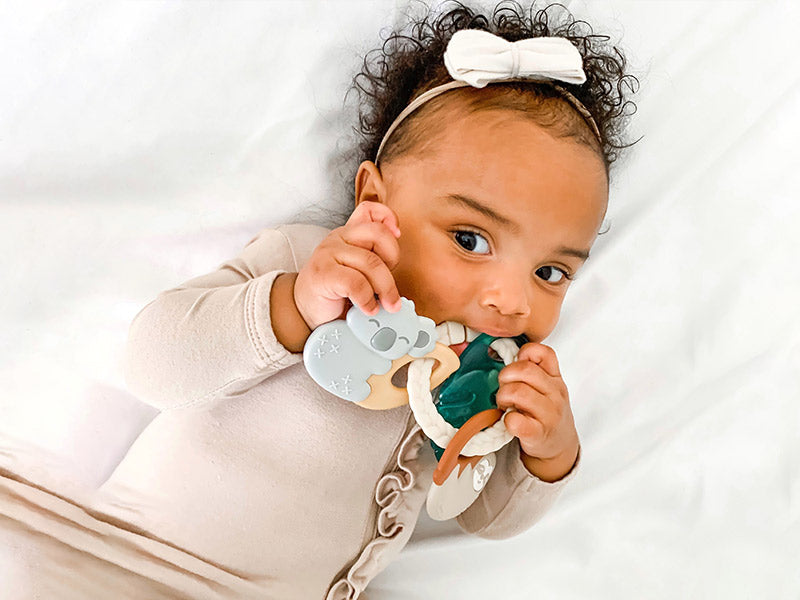 Infant girl with a white bow and cream colored side ruffled long sleeve top sucking on a teething ring. 