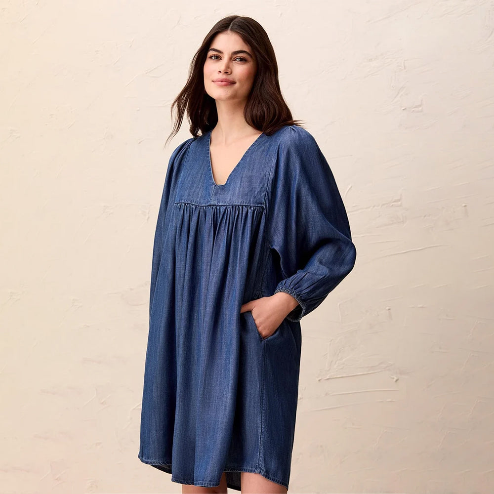 A woman wearing a loose, oversized blue denim tunic dress with a V-neck, smocked chest detail, and long sleeves, standing against a textured beige wall.
