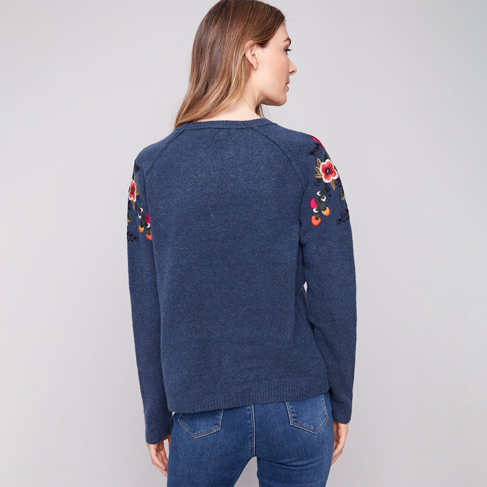 Woman wearing a blue sweater with floral patterns on the sleeves against a gray background