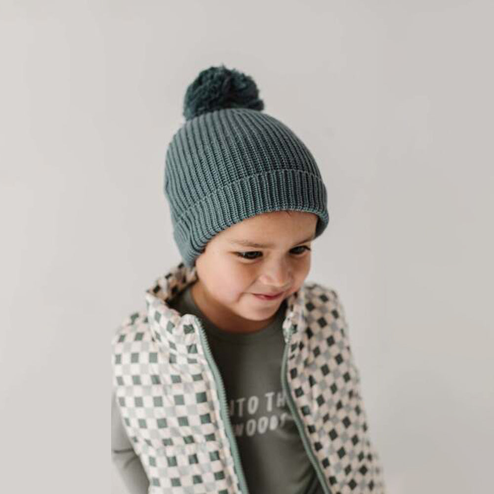 Child wearing a teal knit beanie with a pom-pom on a plain background
