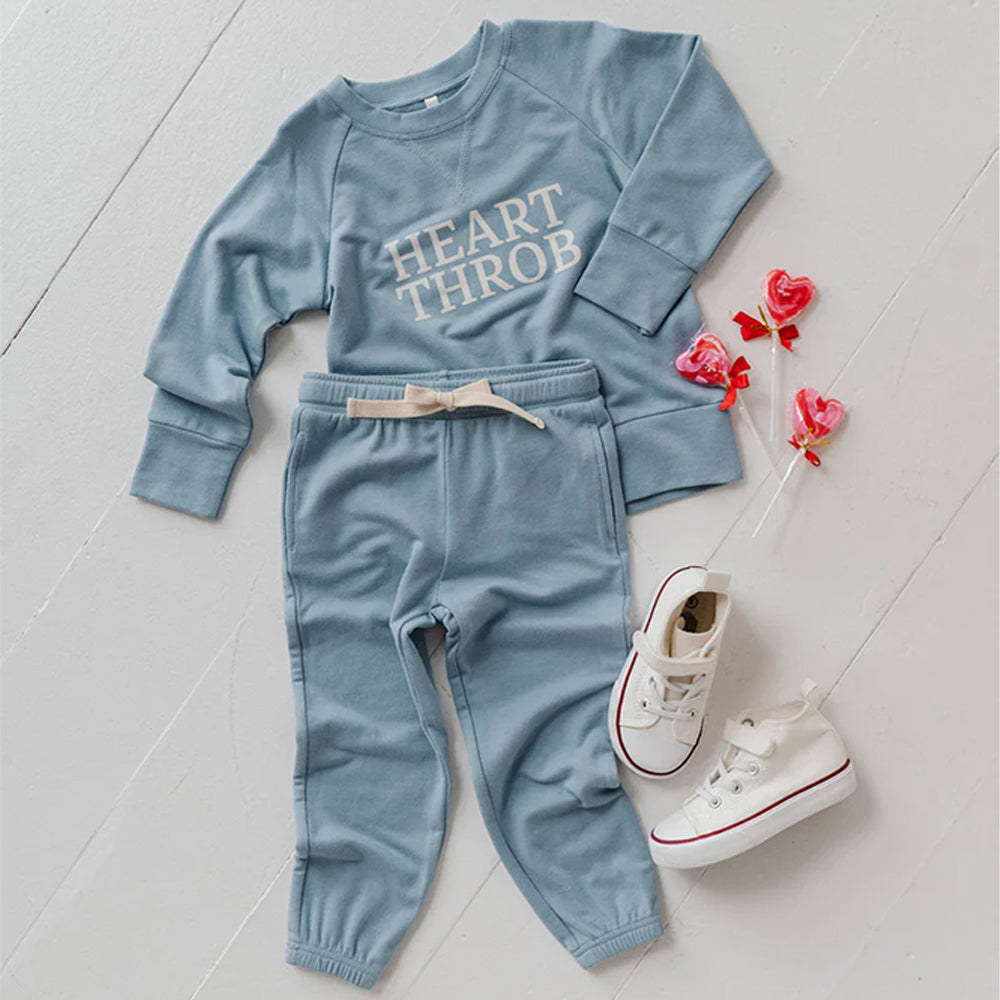 Children's blue outfit with 'HEART THROB' text, white sneakers, and decorative flowers on a light wooden floor.
