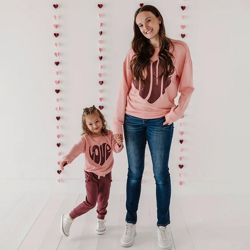 Woman and child wearing matching pink sweatshirts with heart designs against a heart-themed background.