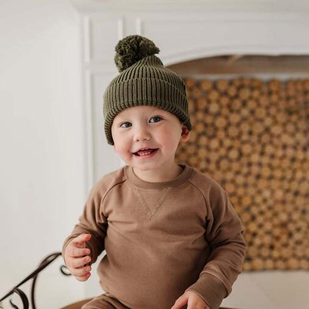 Child wearing a green knit hat with a pom-pom, sitting in front of stacked firewood.