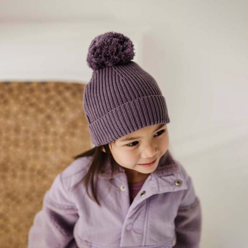 Child wearing a purple knit hat with a pom-pom and matching coat against a neutral background