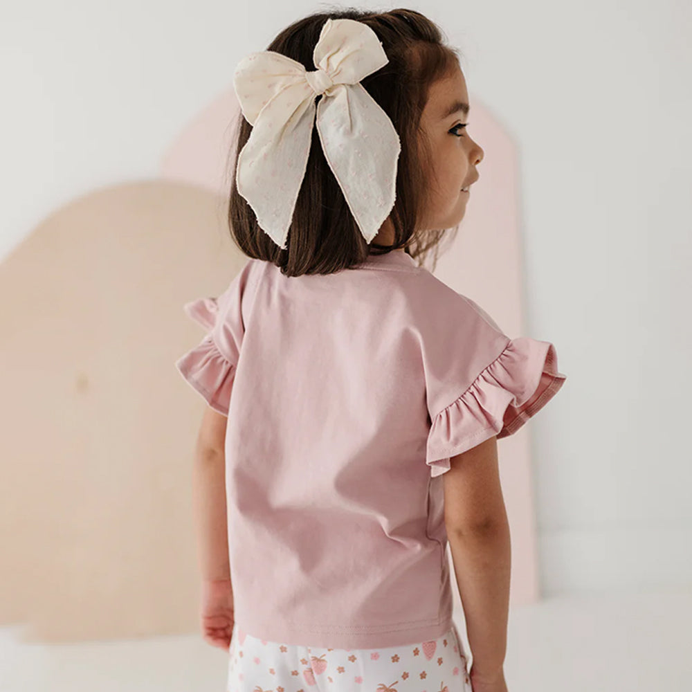 Child wearing a pink top with ruffled sleeves and white pants, standing against a light background.