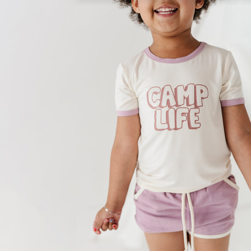 A child wearing a 'Camp Life' t-shirt and purple shorts with a thistle design.