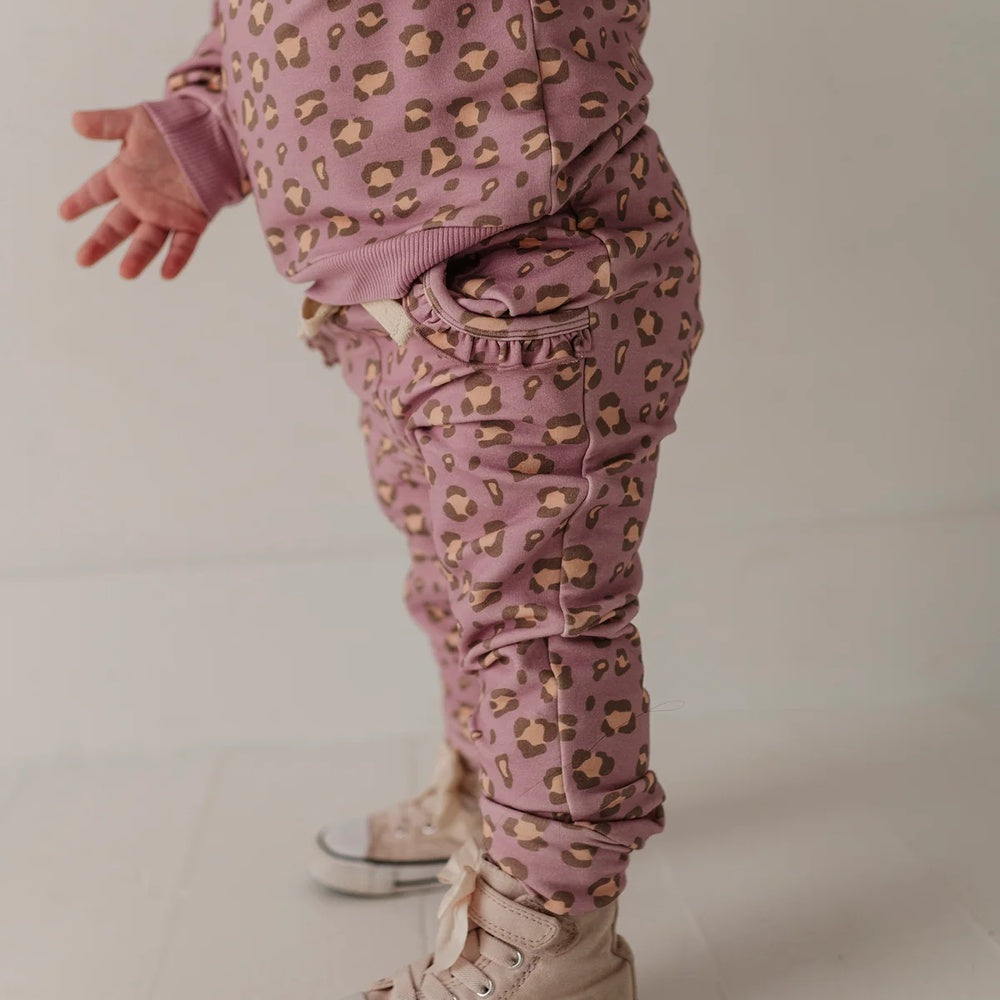 A close-up of a toddler's lower body wearing dusty pink leopard-print pants and a matching top, paired with beige high-top sneakers, against a neutral light gray background.
