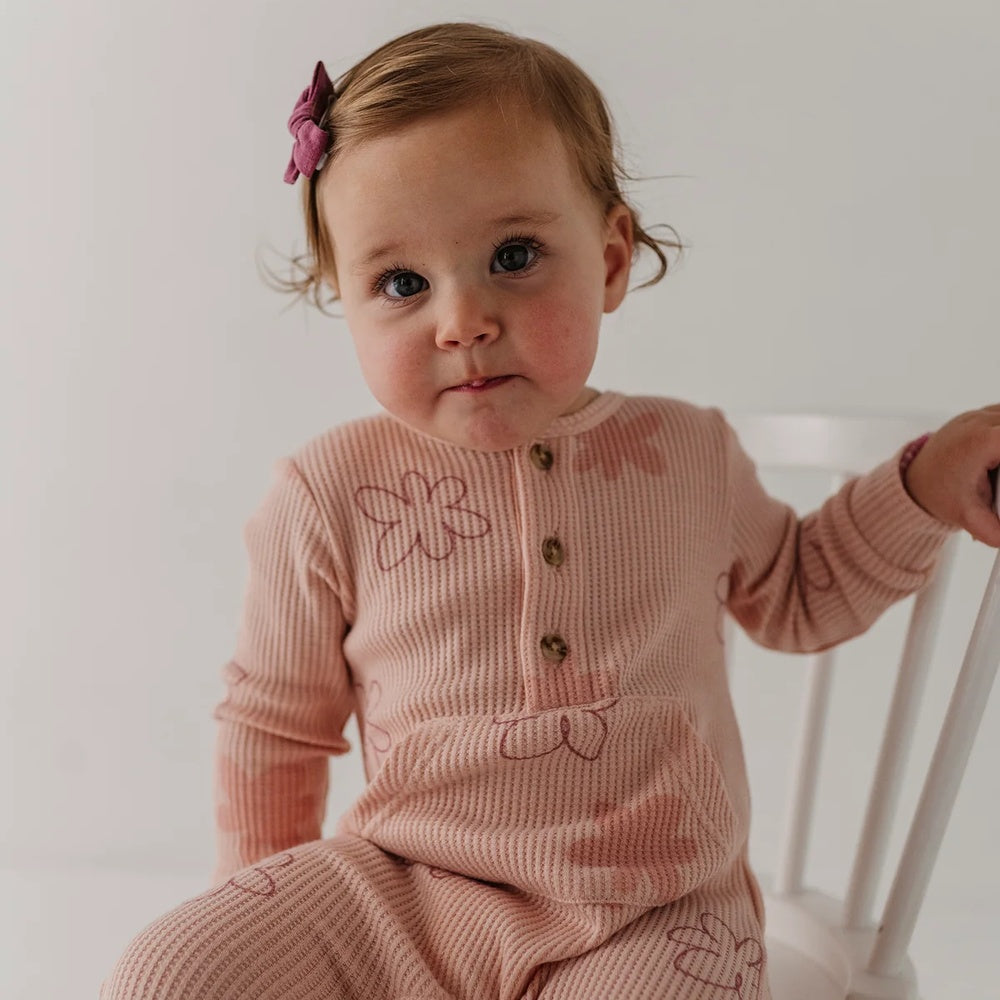 A baby girl wearing a dusty pink ribbed long-sleeve romper with embroidered bows and buttons, sitting on a white wooden chair against a plain white background.