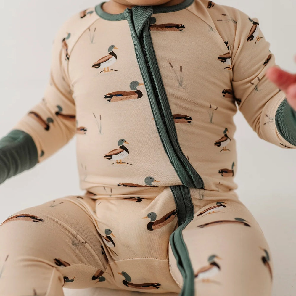 A close-up of a baby wearing a long-sleeve footless sleeper with a duck print on a beige background and dark green trim.