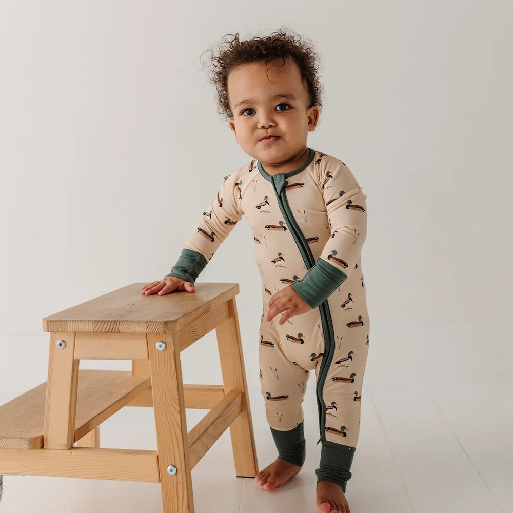 A baby wearing a beige long-sleeve footless sleeper with a green zipper and dark green cuffs, standing next to a small wooden step stool.