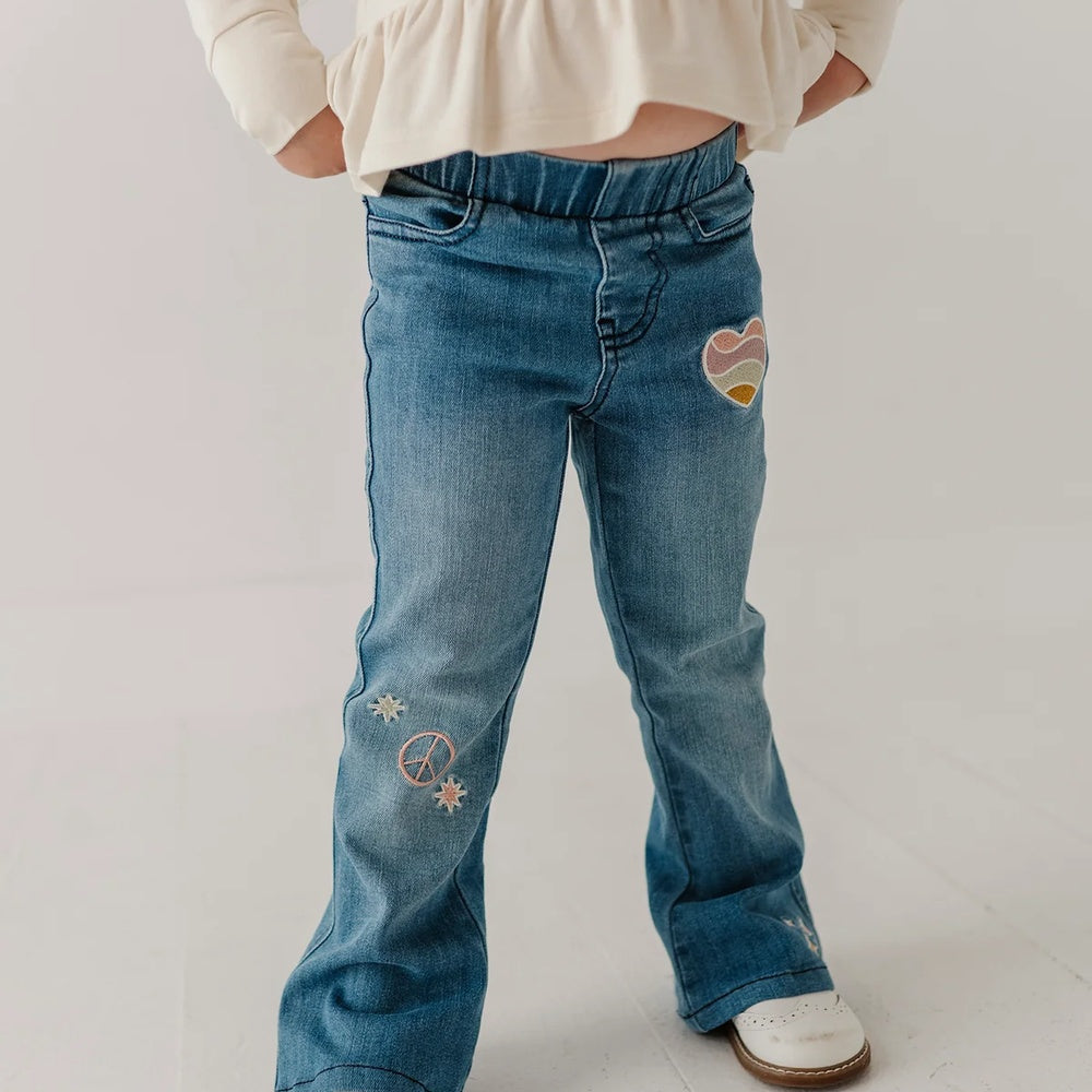 A child wearing blue denim jeans with embroidered patches, including a peace sign and a heart design, paired with a cream long-sleeve top and white shoes.
