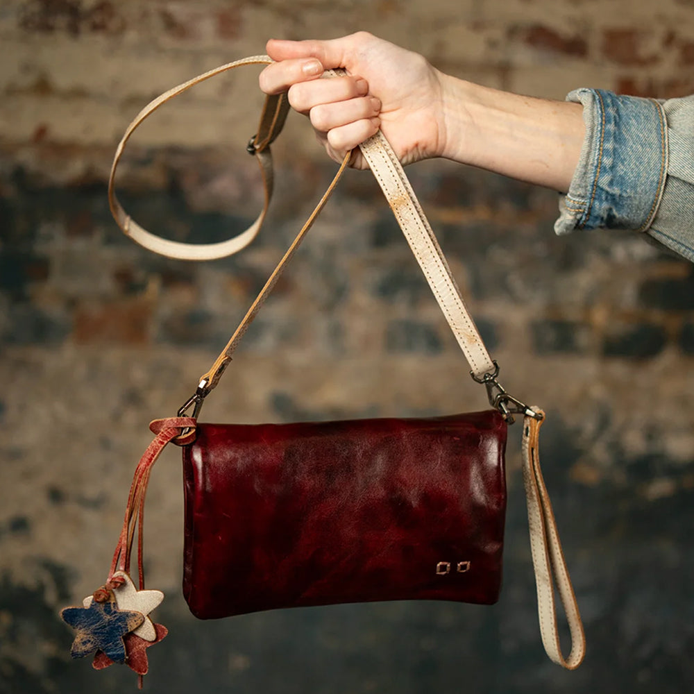 A hand holds a small, dark red leather shoulder bag with a cream-colored strap and leather charms against a rustic, blurred brick wall background.