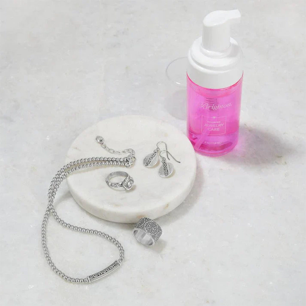 Silver jewelry including a necklace, ring, and earrings on a marble surface with a pink bottle.