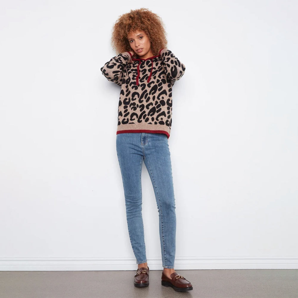 A model with curly brown hair stands against a white wall wearing a beige leopard-print hoodie with red trim, blue skinny jeans, and brown loafers.