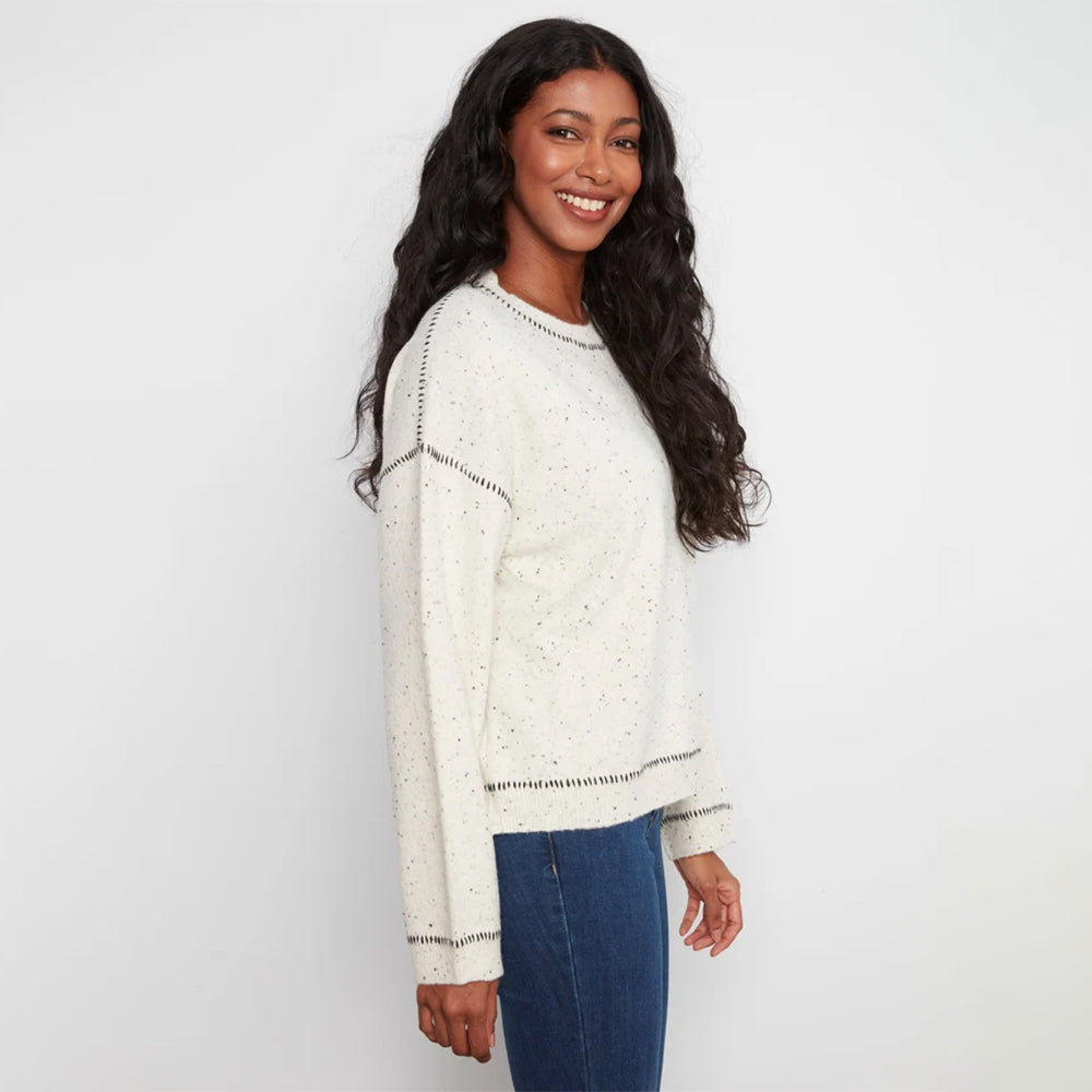 A woman with long, wavy black hair is shown from a side-back angle wearing a cream-colored knit sweater with subtle speckles and dark contrast stitching, paired with blue jeans, against a plain white studio background.