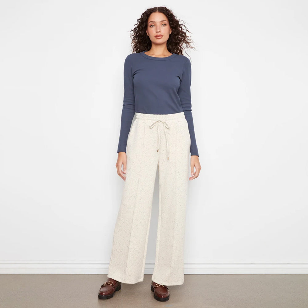 A woman stands against a plain white wall wearing a slate blue long-sleeve top and cream wide-leg drawstring pants, paired with brown loafers.