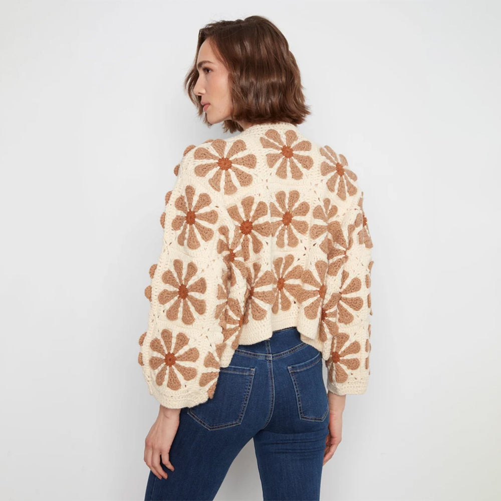 A woman shown from the back wearing a cream crochet cardigan with a brown daisy pattern, paired with blue jeans, against a plain light gray background.
