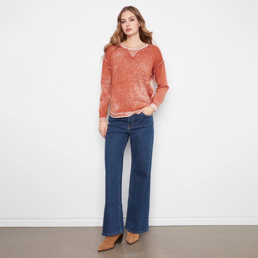 A woman stands against a white wall wearing a rust-orange textured knit sweater with white trim, dark blue wide-leg jeans, and tan ankle boots.