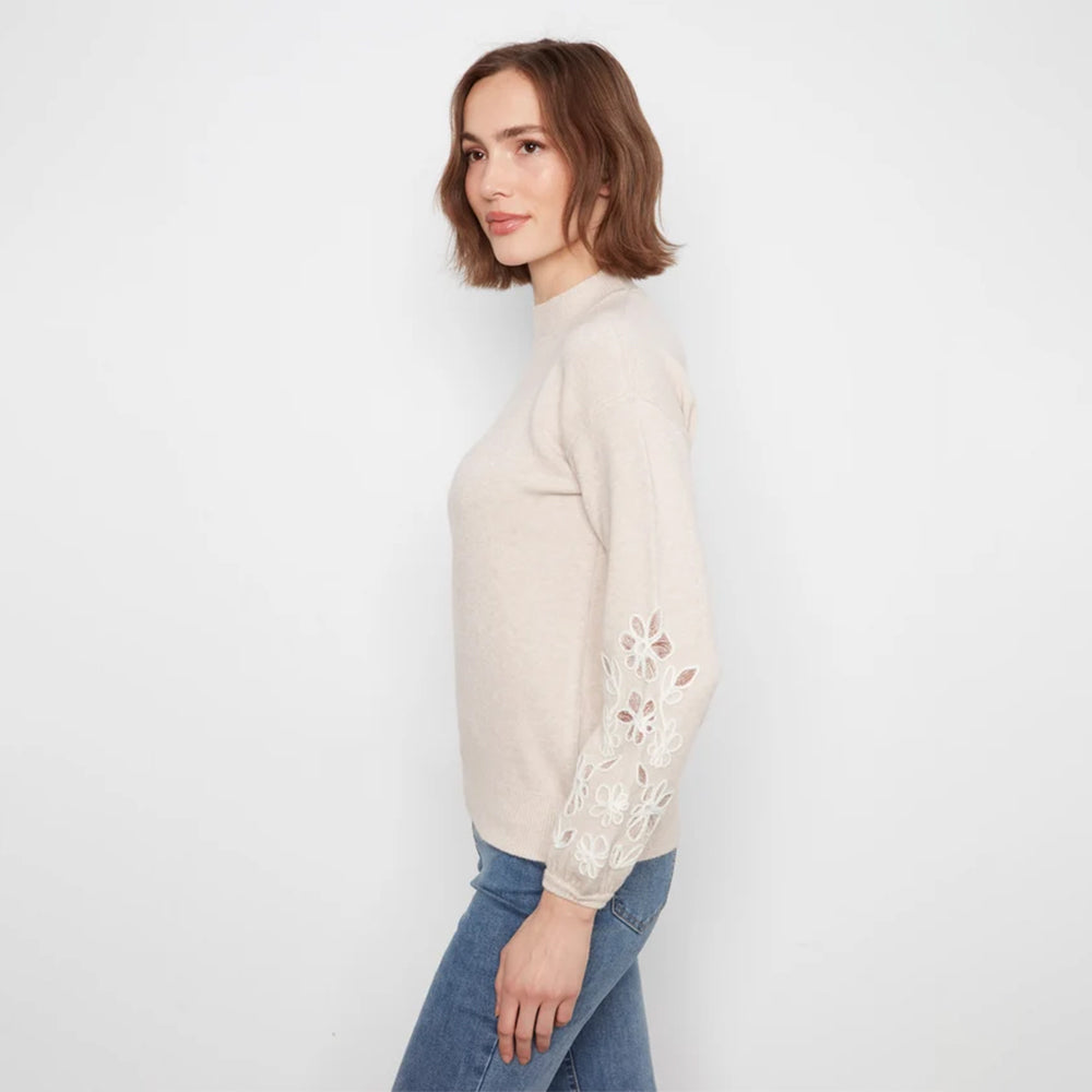 A woman wearing a light beige mock-neck knit sweater with intricate floral lace embroidery on the sleeves, paired with blue jeans, photographed against a clean white studio background.