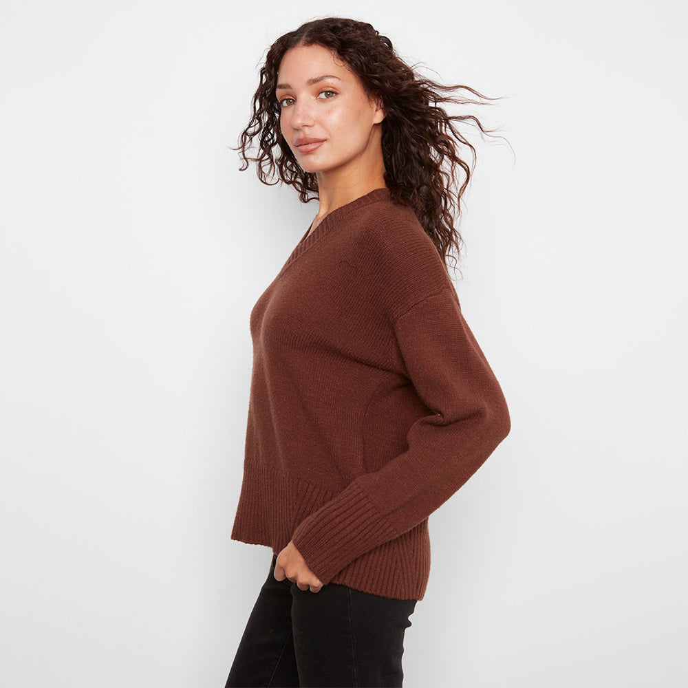 A woman wearing a brown ribbed knit sweater and black pants, photographed from the side against a plain light gray studio background.