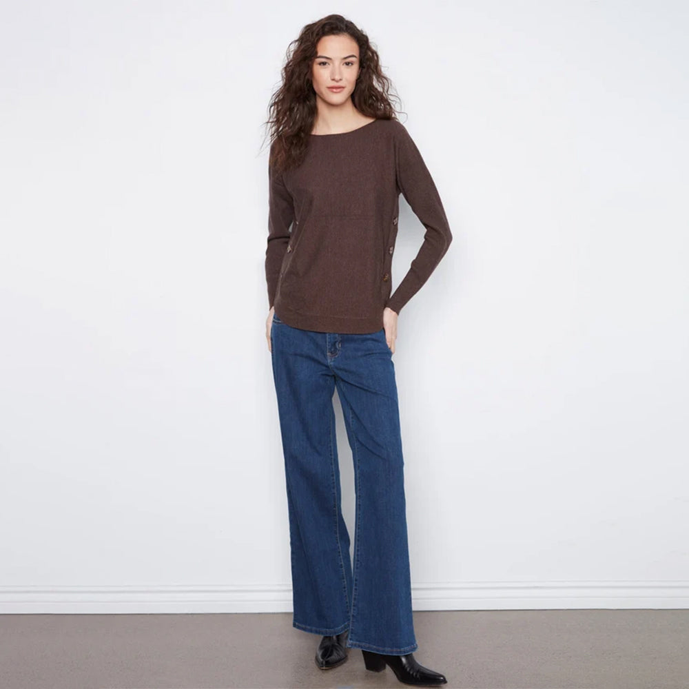 A woman modeling a brown long-sleeve knit top with side pockets and blue wide-leg jeans, standing against a plain white wall.