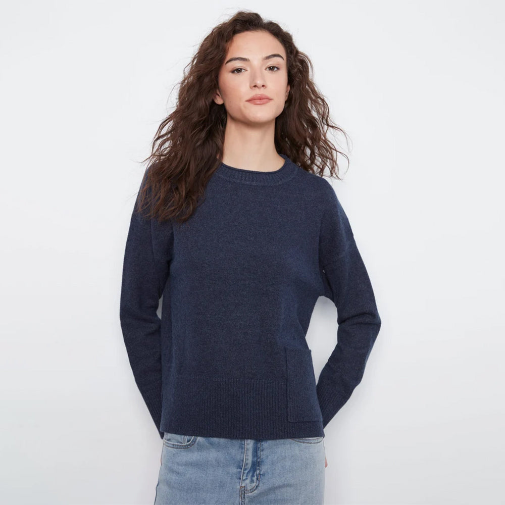 A woman with long, wavy brown hair wearing a dark navy blue crewneck sweater and light blue jeans, standing against a plain white studio background.