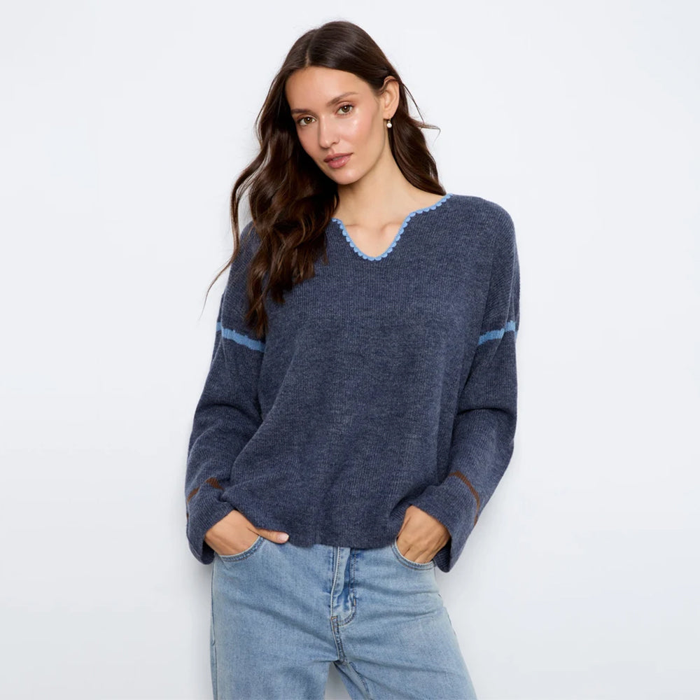 A woman modeling a navy blue knit sweater with light blue trim details on the v-neck and sleeves, paired with light-wash blue jeans against a plain white studio background.
