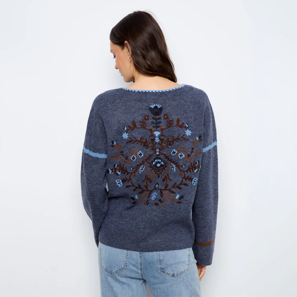 A woman viewed from the back wearing a heathered navy blue knit sweater with a large brown and light blue embroidered snowflake design on the back, paired with light blue jeans, against a plain white background.