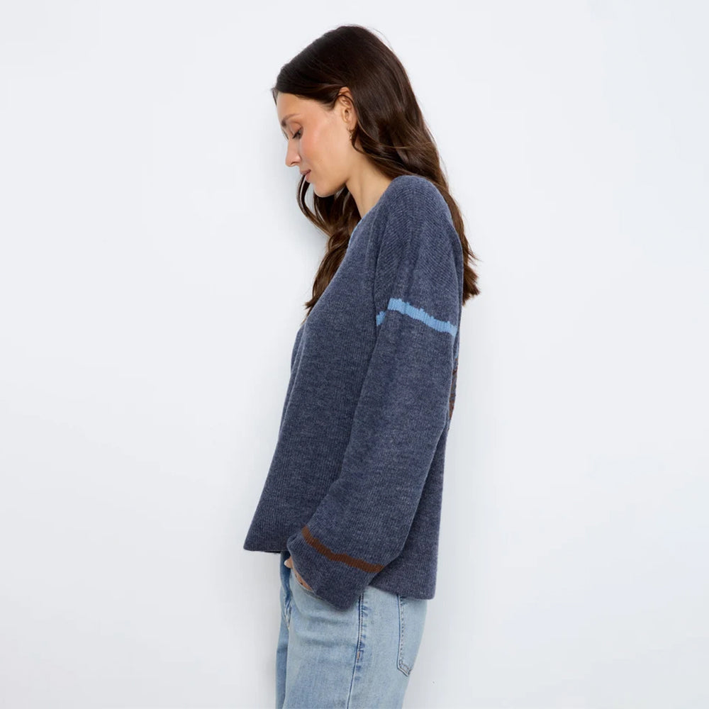 A woman wearing a navy blue knit sweater with light blue and brown stripe accents on the sleeves, paired with light-wash jeans, standing against a plain white wall.