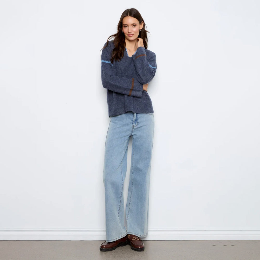 A woman stands against a plain white wall wearing a dark blue-gray knit sweater with light blue wide-leg jeans and brown leather loafers.