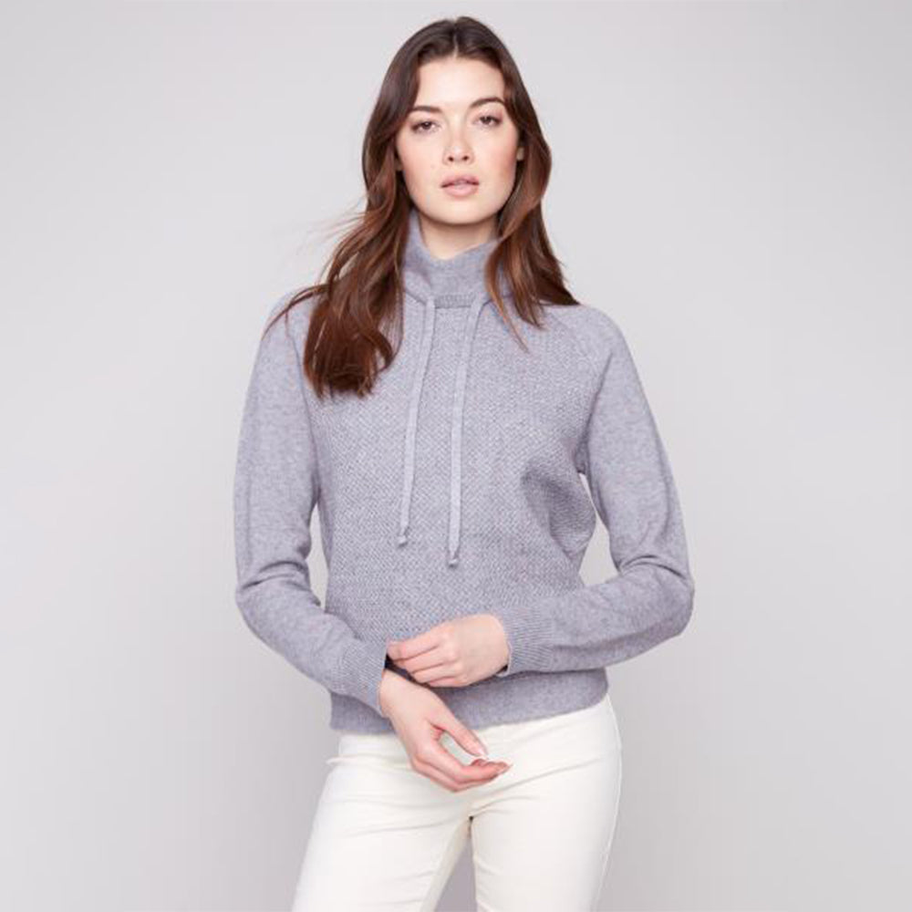 Woman wearing a gray sweater and white pants against a light gray background