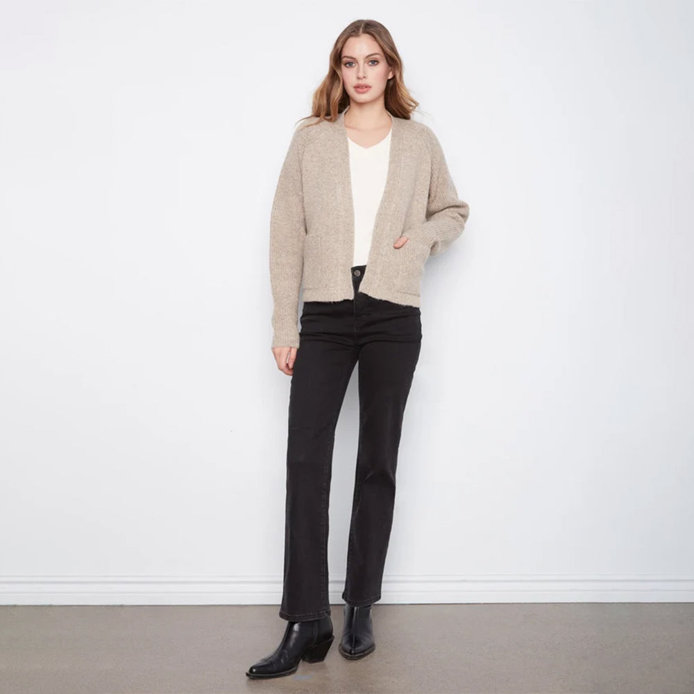 A woman stands against a white wall wearing a light beige knit cardigan over a white top, black straight-leg jeans, and black ankle boots.