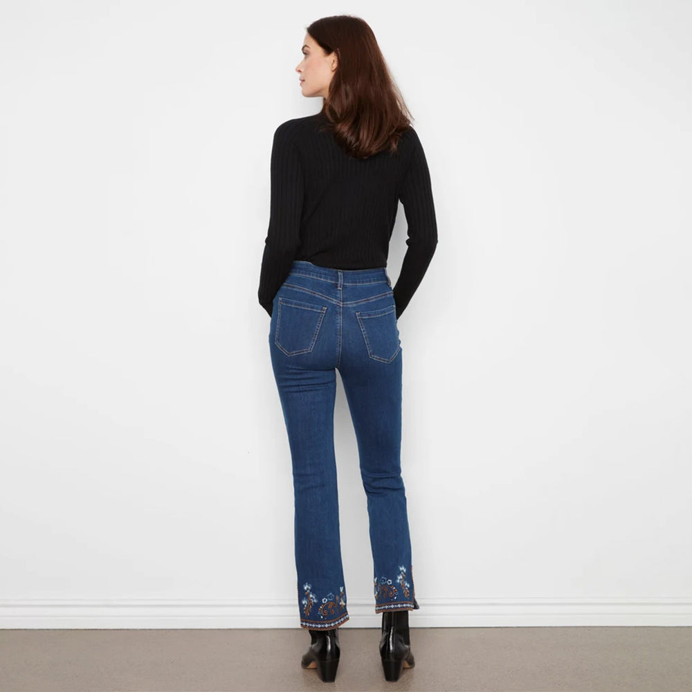 A woman is shown from behind wearing dark blue flare jeans with floral embroidery at the hem, a black ribbed turtleneck sweater, and black ankle boots, standing against a plain white wall.