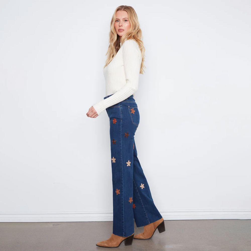 A woman stands in profile wearing blue flare jeans with embroidered star and flower motifs, a white ribbed long-sleeve top, and brown heeled boots against a plain white wall.
