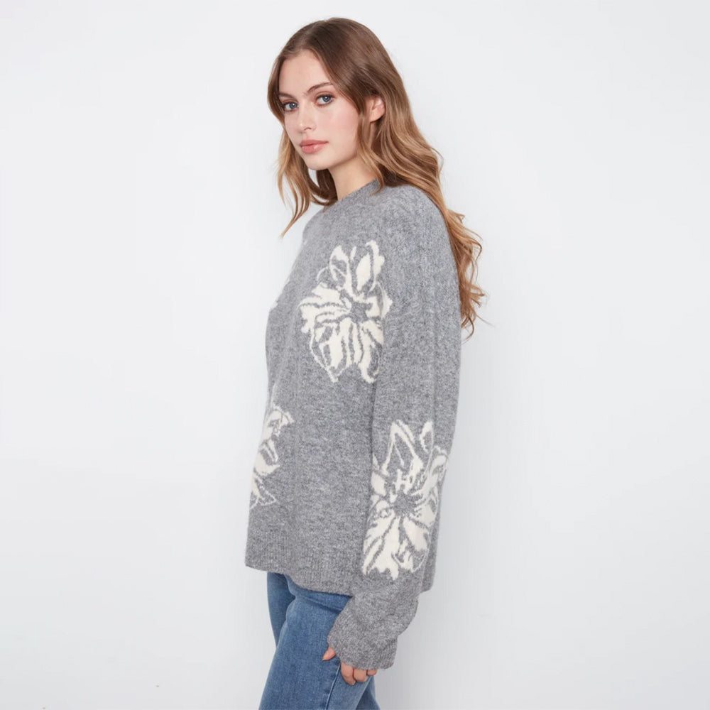 A woman wearing a gray knit sweater with large white floral patterns on the front and sleeves, paired with blue jeans, photographed against a plain white studio background.