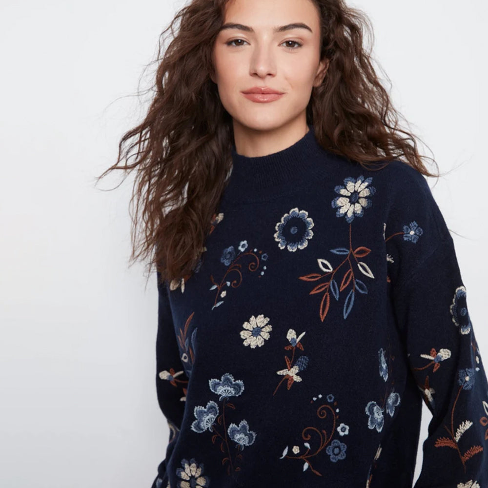 A woman wearing a navy blue mock-neck knit sweater with a delicate embroidered floral pattern in blue, white, and rust orange, set against a plain light gray background.