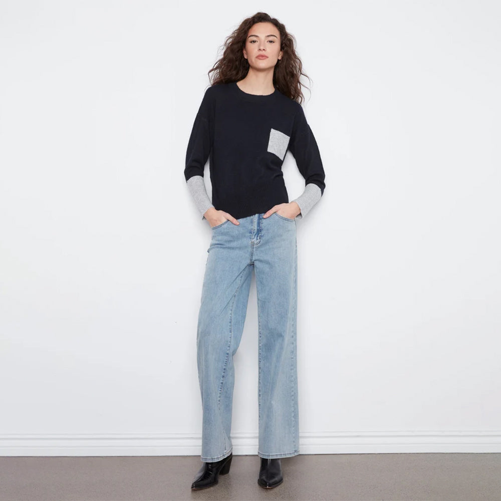 A woman modeling a black long-sleeve knit sweater with a contrasting light gray chest pocket and light gray ribbed cuffs, paired with light blue jeans against a plain white studio background.