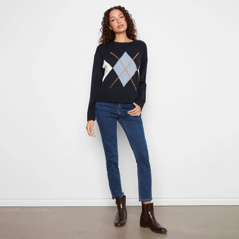 A woman stands against a white wall wearing a dark navy sweater with a light blue and white argyle pattern on the chest, paired with dark blue jeans and dark brown ankle boots.