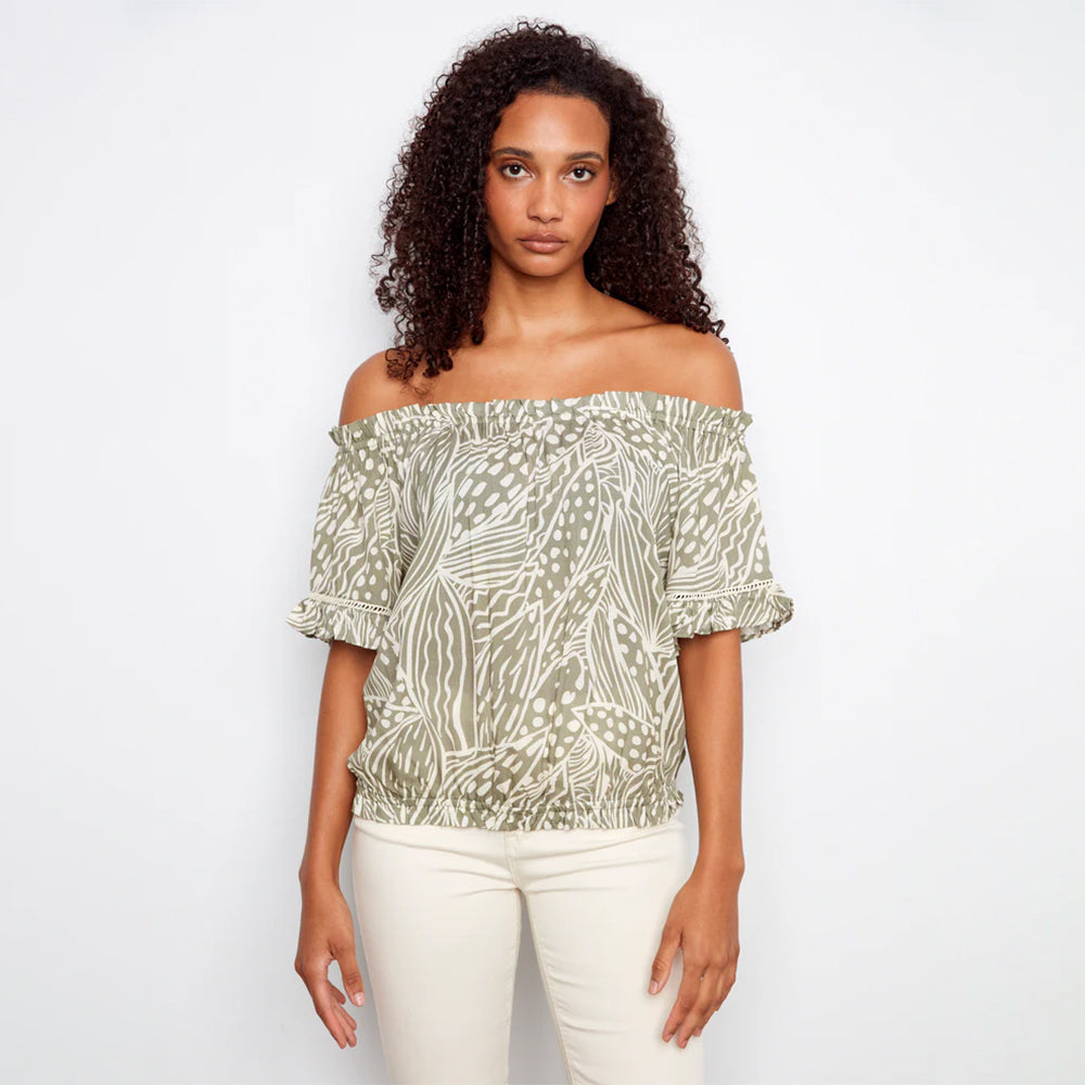 Woman wearing a patterned off-shoulder top and white pants on a plain background