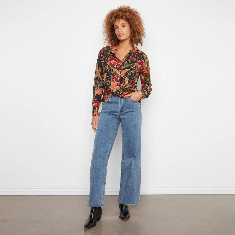 A woman stands against a white wall wearing a long-sleeve button-up blouse with a dense red, orange, and green floral print, paired with blue jeans and black ankle boots.