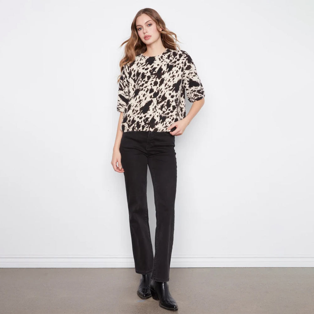A woman stands against a white wall wearing a beige and black abstract-patterned short-sleeve top, black pants, and black ankle boots.