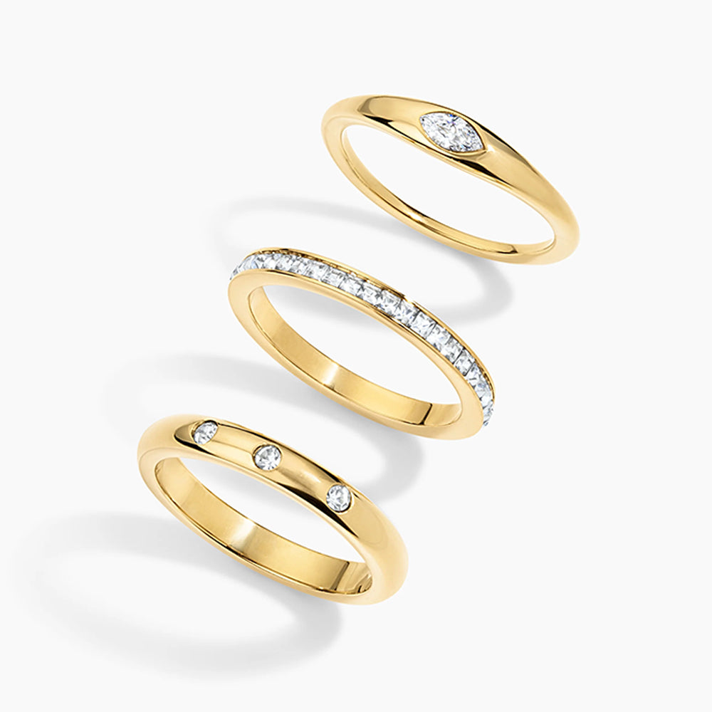 Three yellow gold rings with diamonds, arranged diagonally on a light gray background. One ring features a marquise-cut diamond, one is a pavé band, and one has two small round diamonds.