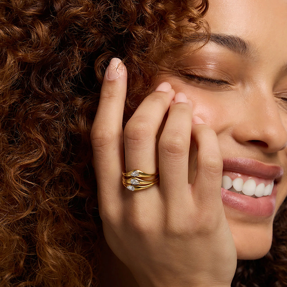 A close-up lifestyle shot of a woman with curly brown hair wearing a stack of three gold rings on her finger. The rings feature clear gemstones, set against a dark background.