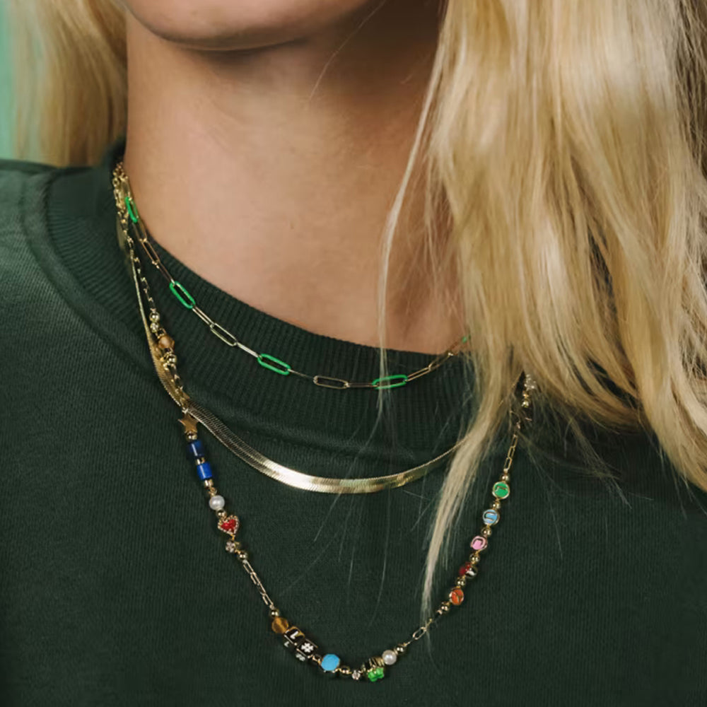 A close-up of a person wearing a layered necklace set. The set includes a gold paperclip chain with green links, a thin gold band, and a longer gold chain decorated with colorful beads and charms.