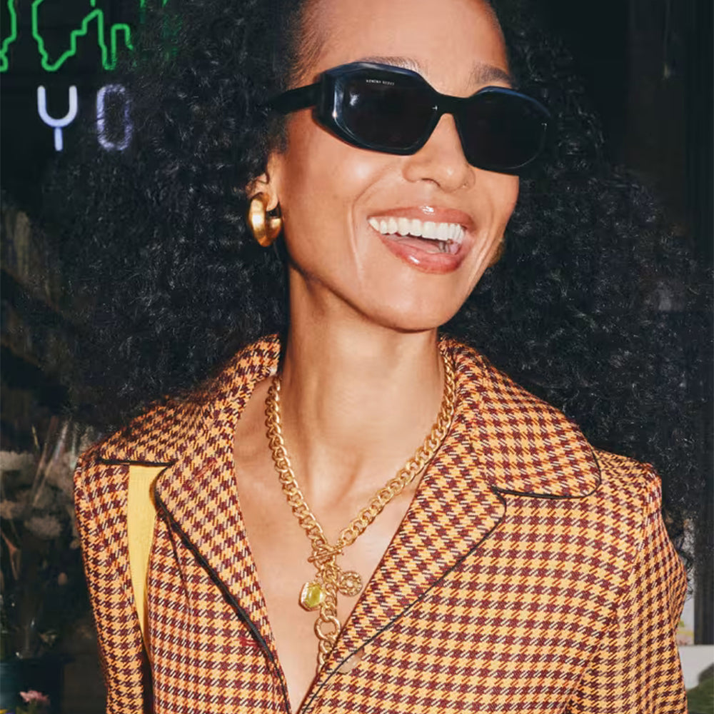A close-up lifestyle shot of a smiling woman with curly hair wearing black sunglasses, a brown and red houndstooth blazer, and gold jewelry.