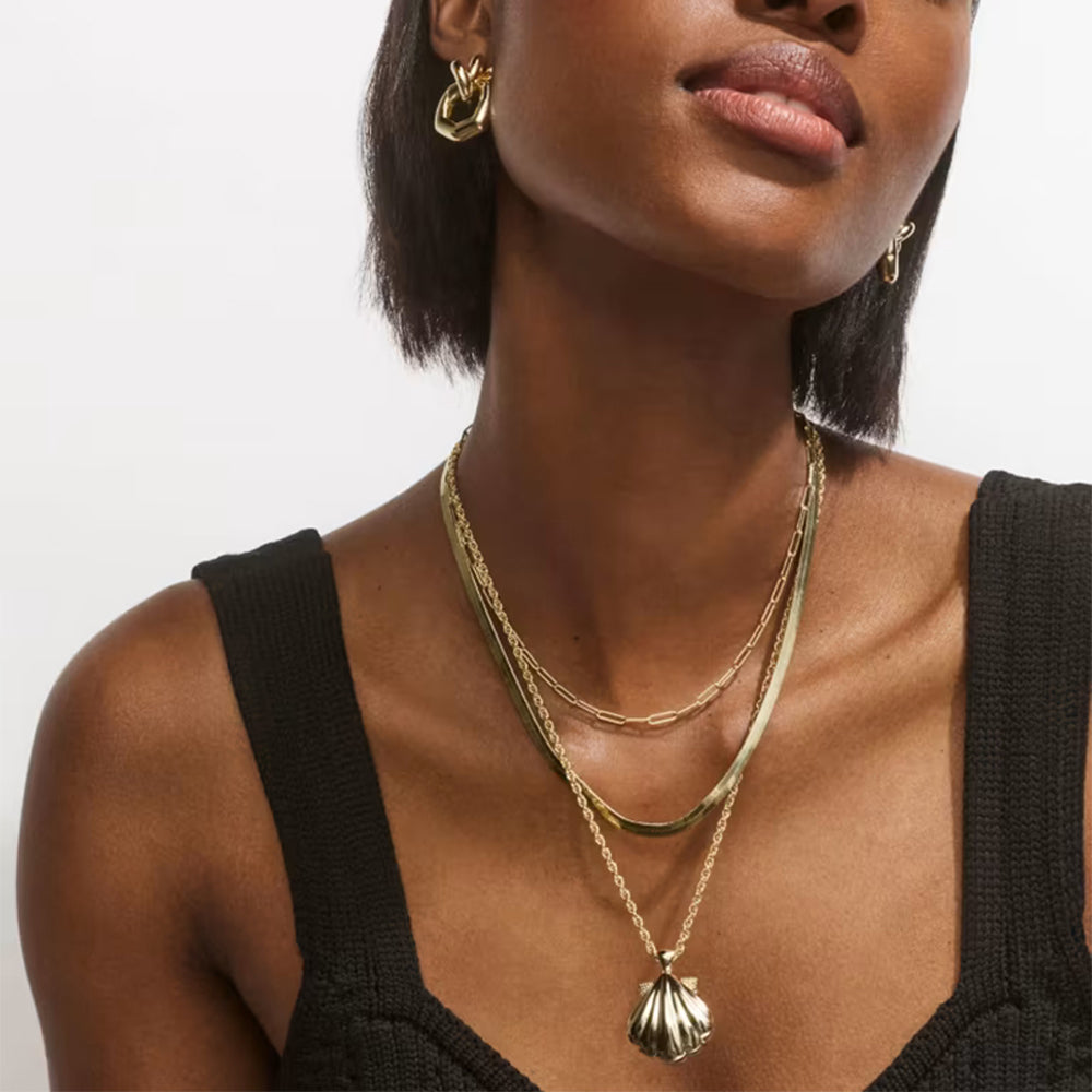 A close-up of a model wearing a layered gold necklace set with a scallop shell pendant, paired with a black knit top against a light gray background.