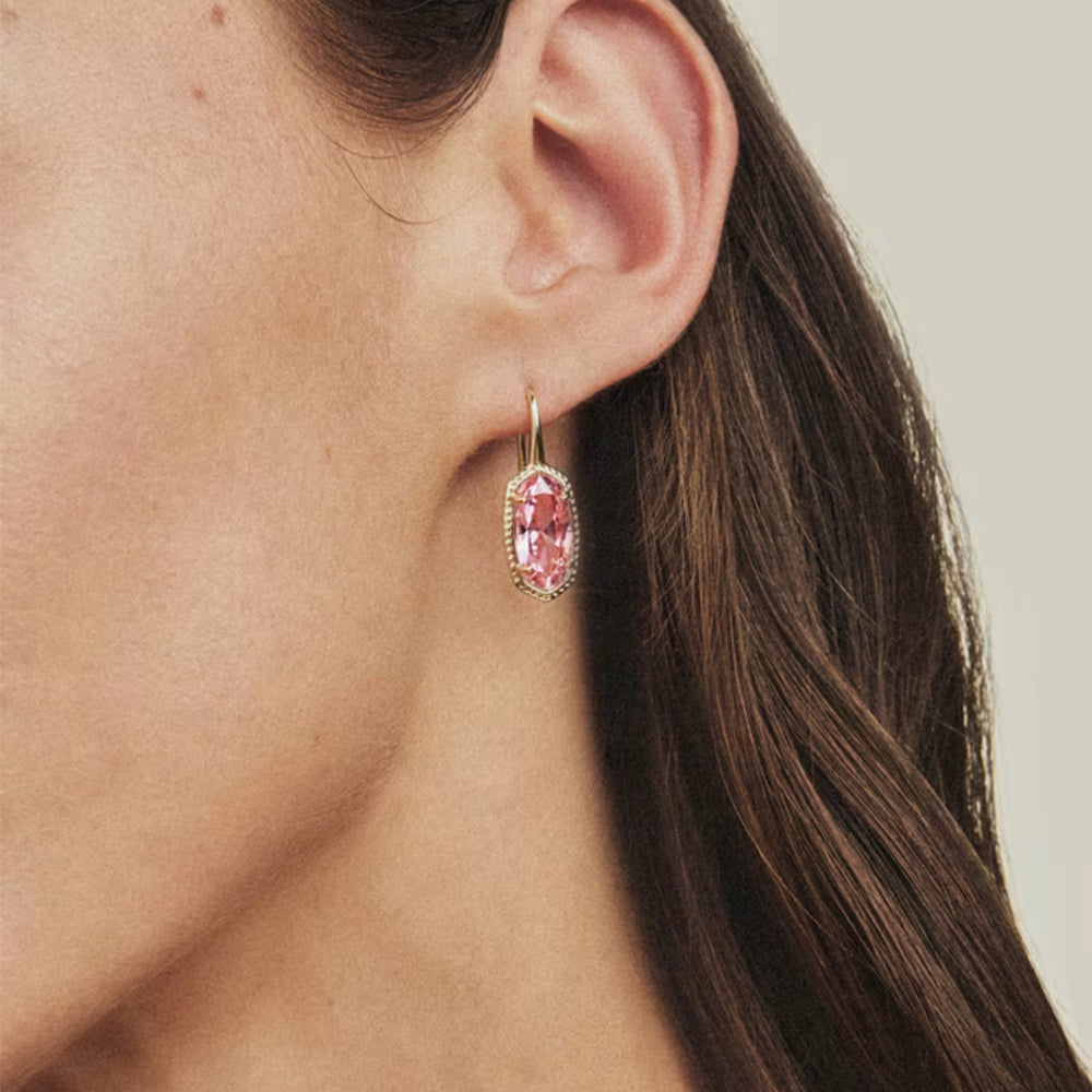 A close-up of a woman's ear wearing a single dangling earring with a marquise-cut pink gemstone in a gold-toned setting with small clear stones around it.