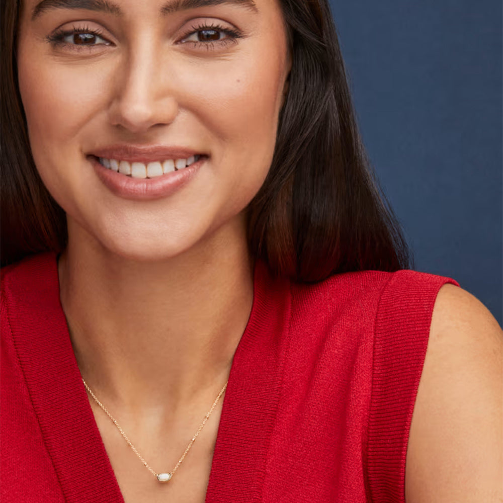 A close-up of a smiling woman wearing a delicate gold necklace with a small white bead pendant, paired with a red ribbed knit top against a dark blue background.