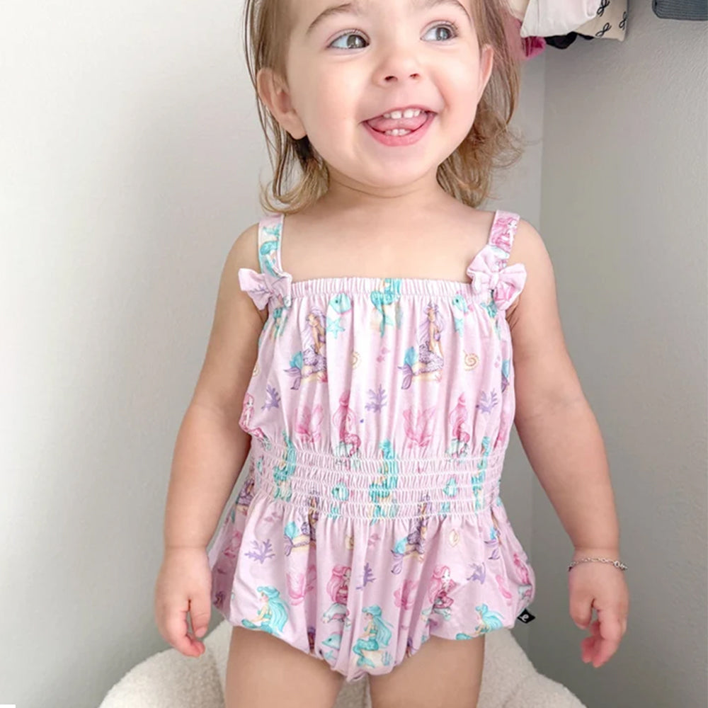 A smiling toddler girl wearing a pink ruffle-strap romper with a mermaid and sea life print, standing indoors against a light gray wall.